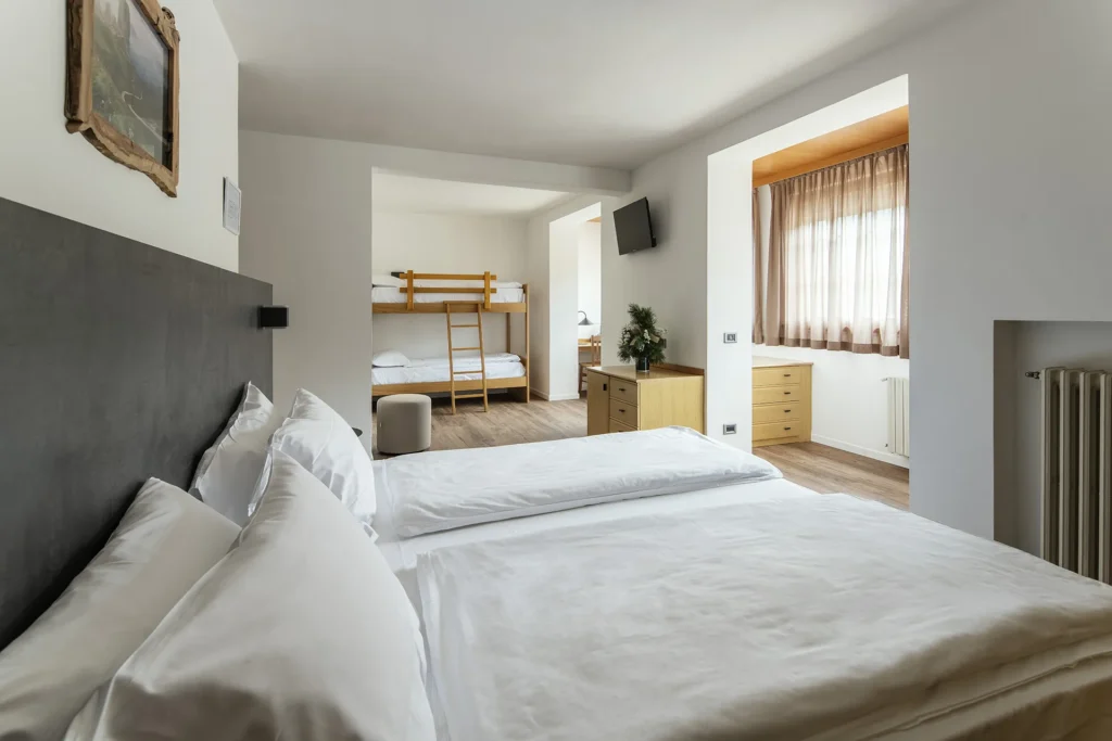 Nell'immagine la camera family dell'hotel con il letto matrimoniale in primo piano con testata grigia. Sulla parete sopra il letto è appeso un quadro e di fronte c'è un termosifone. A destra c'è un'apertura su un altro spazio con una cassettiera e una grande finestra coperta da tende. Il resto della stanza mostra un'altra cassettiera con una tv appesa sopra alla parete e, oltre, un letto a castello. Il pavimento è in parquet.